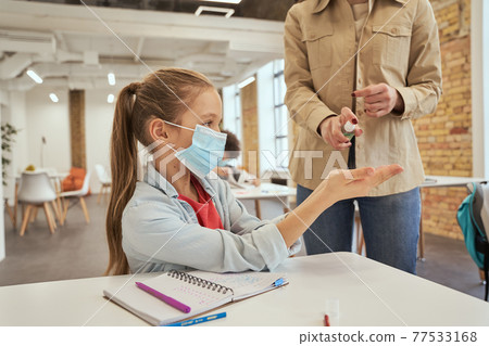 Hygiene first. Young female teacher using an alcohol spray to disinfect student hands in classroom Hygiene first. Young female teacher using an alcohol spray to disinfect student hands in classroom 77533168