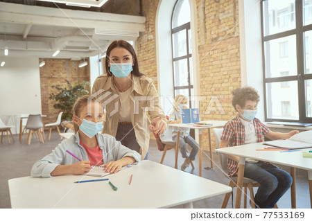 Attractive young female teacher wearing protective face mask helping little girl during lesson. Kids studying in elementary school, sitting at the desk Attractive young female teacher wearing protective face mask helping little girl during lesson. Kids studying in elementary school, sitting at the desk 77533169