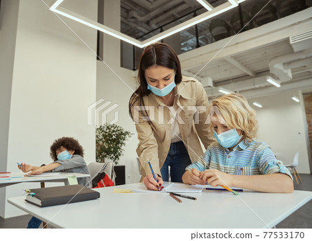 Nice young female teacher wearing protective face mask helping little boy with the task. Kids studying in elementary school, sitting at the desk Nice young female teacher wearing protective face mask helping little boy with the task. Kids studying in elementary school, sitting at the desk 77533170