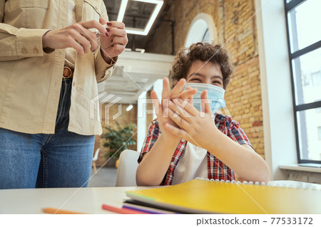 Safety is important. Playful little school boy wearing protective mask cleaning his hands. Female teacher using an alcohol spray to disinfect student hands in classroom 77533172