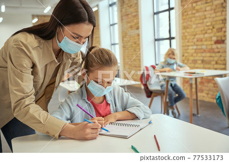 Helpful young female teacher wearing protective face mask helping little girl doing sums. Kids studying in elementary school, sitting at the desk Helpful young female teacher wearing protective face mask helping little girl doing sums. Kids studying in elementary school, sitting at the desk 77533173