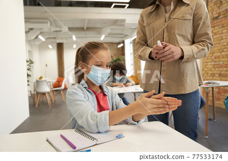 Attractive little school girl wearing protective mask cleaning her hands. Female teacher using an alcohol spray to disinfect student hands in classroom Attractive little school girl wearing protective mask cleaning her hands. Female teacher using an alcohol spray to disinfect student hands in classroom 77533174