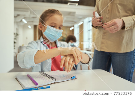 Safety rules. Close up of clean hands of little school girl wearing protective mask. Female teacher using an alcohol spray to disinfect student hands in classroom Safety rules. Close up of clean hands of little school girl wearing protective mask. Female teacher using an alcohol spray to disinfect student hands in classroom 77533176