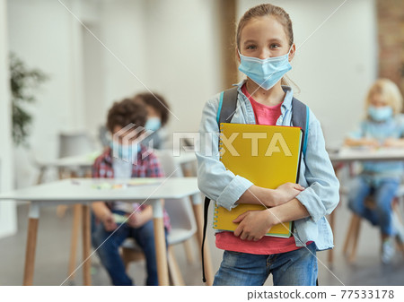 Portrait of happy little school girl wearing mask to prevent the spread of Covid19 looking at camera, holding textbooks while posing in a classroom Portrait of happy little school girl wearing mask to prevent the spread of Covid19 looking at camera, holding textbooks while posing in a classroom 77533178