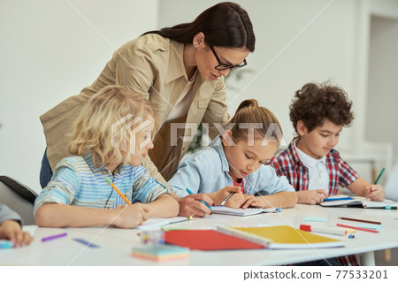 Excellence. Kind young female teacher in glasses helping her little schoolchildren in a classroom. Kids sitting at the table, studying in elementary school 77533201