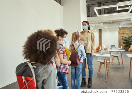 Young female teacher screening school children for fever against the spread of Covid19, using digital thermometer while kids coming to school 77533236