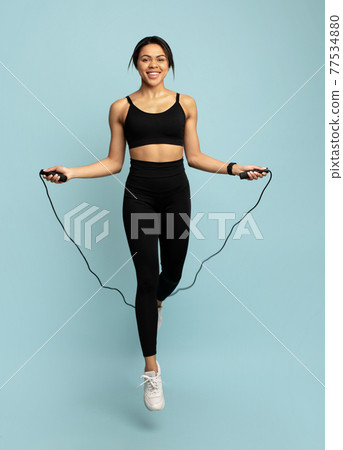 Happy fit african american lady jumping over the skipping rope, during cardio workout on blue background in studio Happy fit african american lady jumping over the skipping rope, during cardio workout on blue background in studio 77534880