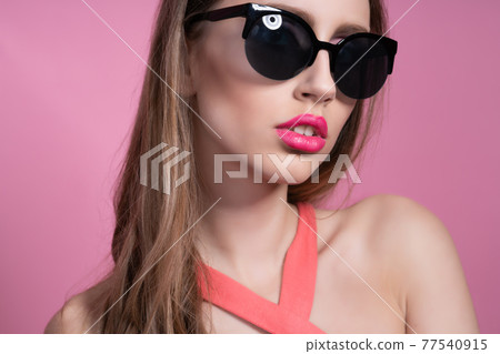 Fashion photo of a beautiful elegant young woman in a pretty pink suit and sunglasses, posing over pink background. 77540915