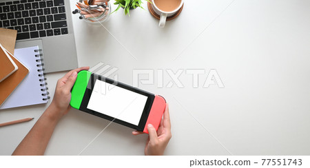 Top view image of hands holding a portable video game with white blank screen over white working desk. Top view image of hands holding a portable video game with white blank screen over white working desk. 77551743