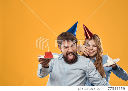 Birthday party man and woman on a yellow background in hats with a cake in their hands Birthday party man and woman on a yellow background in hats with a cake in their hands 77553359