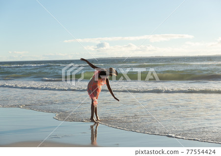 Mixed race woman wearing straw hat walking along the seashore with spread hands 77554424