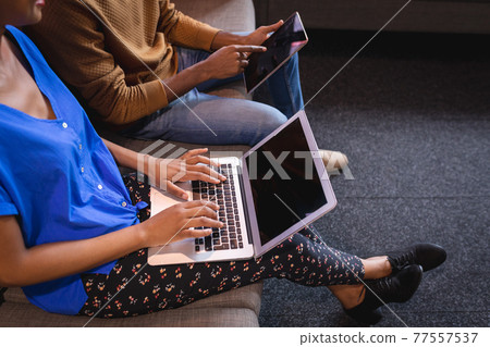 Diverse male and female colleagues working sititng on sofa in office using computer and tablet Diverse male and female colleagues working sititng on sofa in office using computer and tablet 77557537