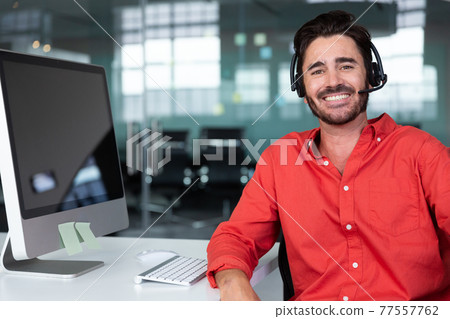 Portrait of caucasian businessman sitting at desk with blank screen wearing headset 77557762