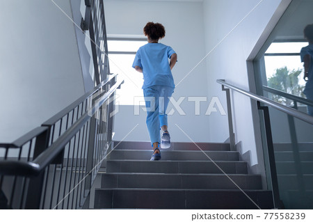 Rear view of mixed race female doctor wearing scrubs running up stairs in hospital 77558239