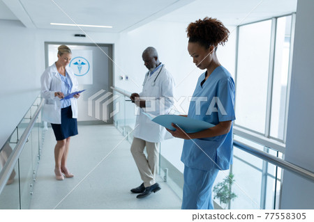 Three diverse male and female doctors standing in hospital corridor looking at medical documentation 77558305