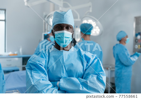 African american male surgeon with face mask and protective clothing in operating theatre 77558661