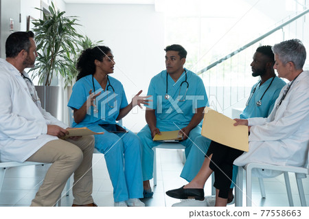 Diverse male and female doctors sitting in hospital corridor and discussing Diverse male and female doctors sitting in hospital corridor and discussing 77558663