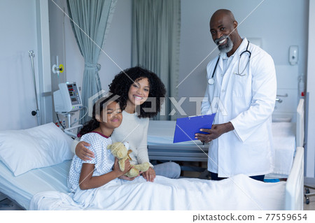 Smiling african american male doctor with mixed race mother and her sick daughter, in hospital bed 77559654