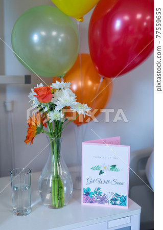 Flowers in vase, get well card and balloons on bedside table in children's ward at a hospital 77559655
