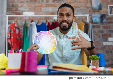 Mixed race male designer in sitting at desk holding a colour chart and smiling Mixed race male designer in sitting at desk holding a colour chart and smiling 77560300