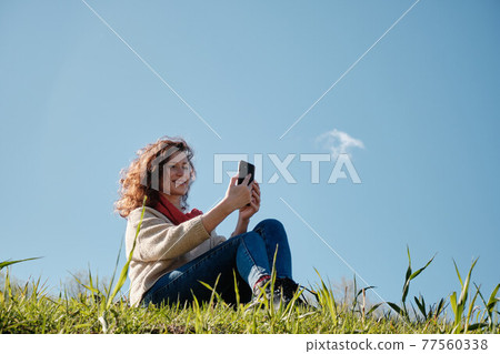 Young girl with a phone in her hands, green grass and blue sky Young girl with a phone in her hands, green grass and blue sky 77560338