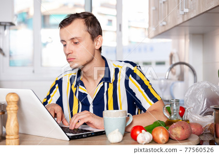 Young man reads a recipe for dishes in a laptop Young man reads a recipe for dishes in a laptop 77560526