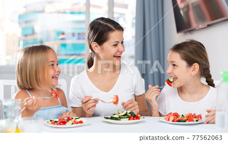 Two funny girls with mom eat vegetable salad at dinner table Two funny girls with mom eat vegetable salad at dinner table 77562063