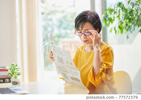 A middle-aged woman wearing reading glasses and reading an English newspaper A middle-aged woman wearing reading glasses and reading an English newspaper 77572379