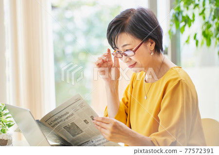 A middle-aged woman wearing reading glasses and reading an English newspaper A middle-aged woman wearing reading glasses and reading an English newspaper 77572391