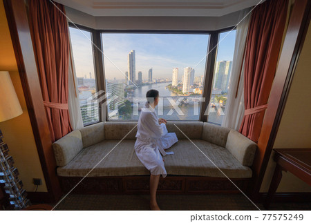 Back of Asian man sitting on sofa couch with window glass in hotel room with Bangkok City skyline in Thailand. Skyscraper buildings. Downtown skyline. Urban town. Relaxing area. People lifestyle. 77575249
