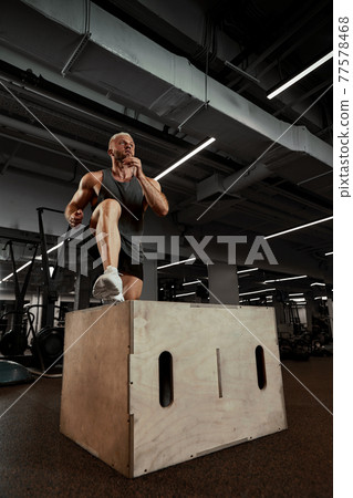 Sexy muscular men using the platform for his legs on a dark colorful background of gym. Sexy muscular men using the platform for his legs on a dark colorful background of gym. 77578468