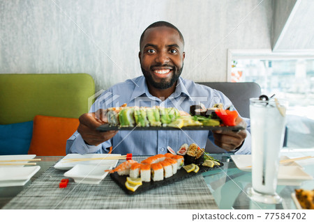 Young african business man sitting at sushi bar, demonstrating to the camera big plate with set of sushi rolls, grinning with cheerful face expression. 77584702