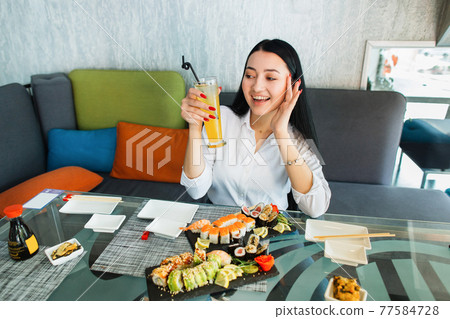 Young brunette Asian woman sitting in cafe at glass table, drinking fresh juice and enjoying her tasty lunch with sushi rolls at modern oriental cafe. Young brunette Asian woman sitting in cafe at glass table, drinking fresh juice and enjoying her tasty lunch with sushi rolls at modern oriental cafe. 77584728