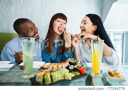 Sushi concept, seafood, asian meal. Three young multiethnic friends, Caucasian and Asian girls, African man, sitting in restaurant, spending funny time with plate of delicious sushi. Sushi concept, seafood, asian meal. Three young multiethnic friends, Caucasian and Asian girls, African man, sitting in restaurant, spending funny time with plate of delicious sushi. 77584778