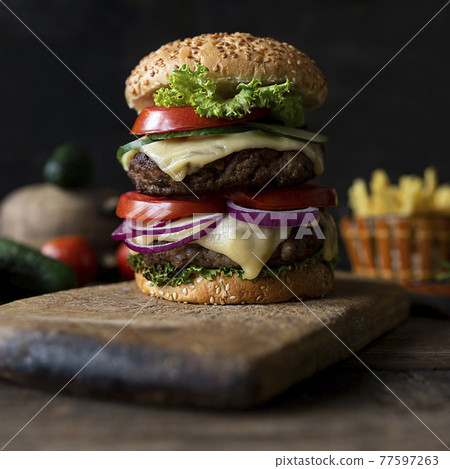 Hamburger with tomatoes, onions, cucumber, lettuce and melting cheese served on a  rustic wooden board with french fries and vegetables in background 77597263
