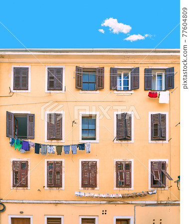 Picturesque typical Italian slum residential house under a blue summer sky with a single cloud in Italy. Wall with many windows with shutters and linen being dried on ropes. Picturesque typical Italian slum residential house under a blue summer sky with a single cloud in Italy. Wall with many windows with shutters and linen being dried on ropes. 77604809