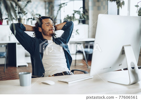 Young businessman sitting in office with hands behind his head satisfied with work done 77606561
