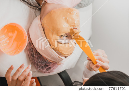 Using a gold mask in cosmetology, a woman at a beautician applying a gold mask on her face. 77611701