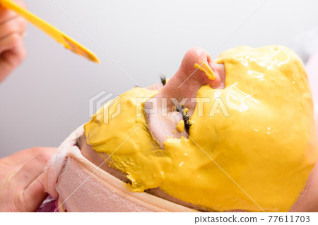 Using a gold mask in cosmetology, a woman at a beautician applying a gold mask on her face. 77611703