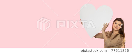 Girl with a large white heart shape in the hands. A student in love stands and holds a large heart shape in her hands. Valentine's Day and love. Panoramic frame. Girl with a large white heart shape in the hands. A student in love stands and holds a large heart shape in her hands. Valentine's Day and love. Panoramic frame. 77611836