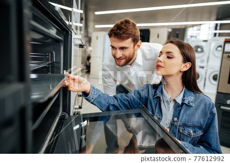 Young couple choosing new electric oven in hypermarket Young couple choosing new electric oven in hypermarket 77612992