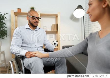 Pregnant woman holding a hand of her disabled husband sitting in a wheelchair Pregnant woman holding a hand of her disabled husband sitting in a wheelchair 77612993