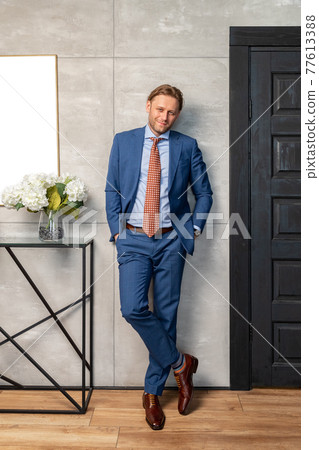 The handsome stylish young man stands at a wall, long curly hair, he is dressed in a expensive blue suit and trousers, he is looking at the camera, a brown classical shoes, Flowers on a curbstone 77613388