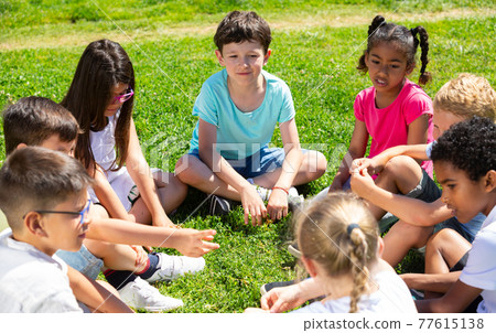 Smiling children friends sitting on grass together and chatting 77615138