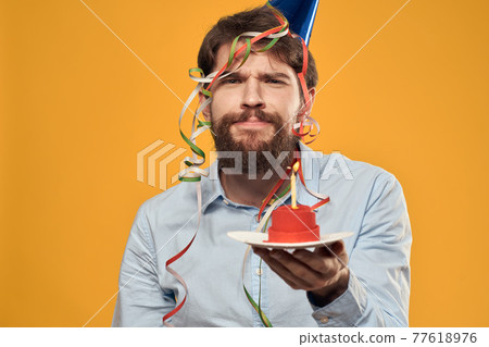 Cheerful man with a cake on a yellow background birthday holidays cap on his head Cheerful man with a cake on a yellow background birthday holidays cap on his head 77618976