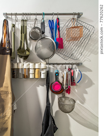 Metal rail with kitchen tools, kitchen utensils hanging, apron and seasoning in front of a white wall in the kitchen. Metal rail with kitchen tools, kitchen utensils hanging, apron and seasoning in front of a white wall in the kitchen. 77620262