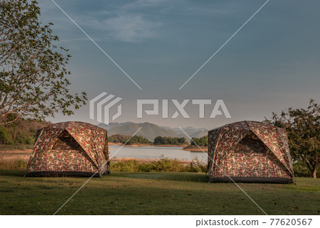 View of two camping tent with awning pitched on the dry ground Near beautiful big trees with branches at the campsite in lake shore with mountain range in background. View of two camping tent with awning pitched on the dry ground Near beautiful big trees with branches at the campsite in lake shore with mountain range in background. 77620567