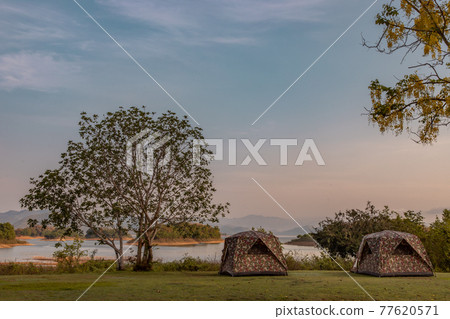 View of two camping tent with awning pitched on the dry ground Near beautiful big trees with branches at the campsite in lake shore with mountain range in background. View of two camping tent with awning pitched on the dry ground Near beautiful big trees with branches at the campsite in lake shore with mountain range in background. 77620571