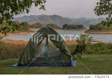View of a camping tent on the dry ground Near beautiful big tree at the campsite in lake shore with mountain range in background. View of a camping tent on the dry ground Near beautiful big tree at the campsite in lake shore with mountain range in background. 77620574