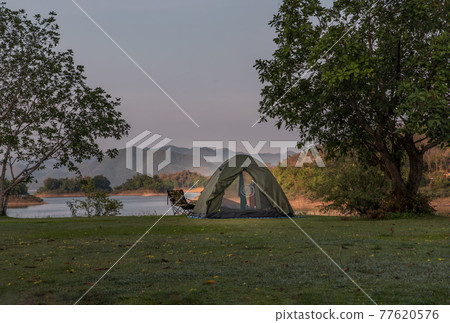 View of a camping tent on the dry ground Near beautiful big tree at the campsite in lake shore with mountain range in background. View of a camping tent on the dry ground Near beautiful big tree at the campsite in lake shore with mountain range in background. 77620576